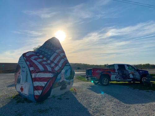 A large painted rock featuring an American flag and a truck parked nearby under a bright sun.