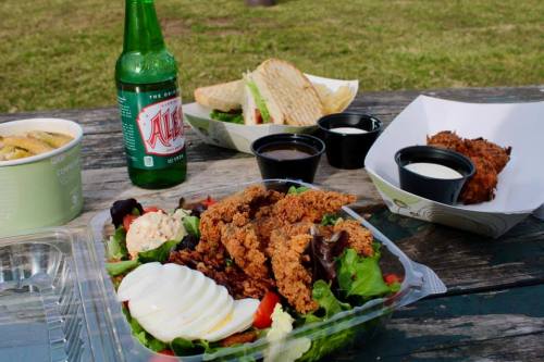 A picnic spread featuring fried chicken salad, sandwiches, dipping sauces, and a green soda on a wooden table.