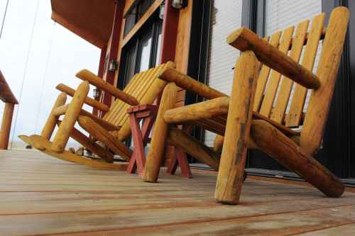 Two rustic wooden rocking chairs and a small table on a wooden deck, with a cozy cabin in the background.