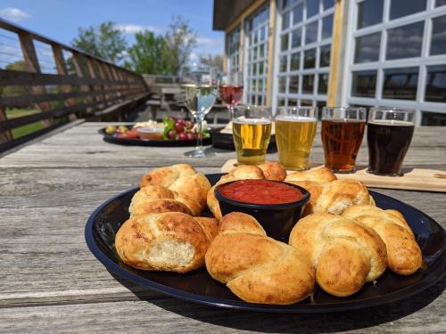 A platter of golden breadsticks with marinara sauce, accompanied by various drinks on a wooden table outdoors.