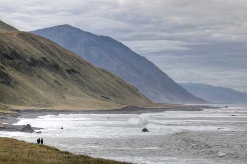 Two people walk along a rocky beach with rolling waves, framed by green hills and distant mountains under a cloudy sky.