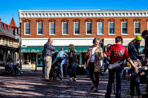 A lively street scene with people socializing and dancing in front of brick buildings under a clear blue sky.