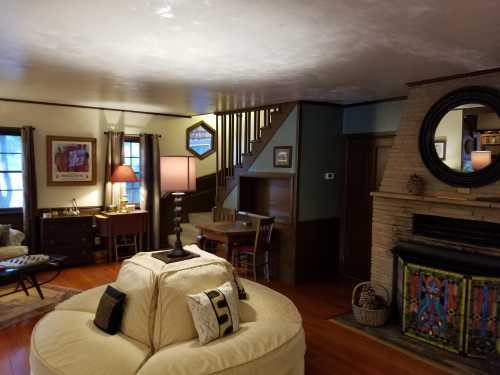 Cozy living room with a circular sofa, lamp, staircase, and a colorful fireplace, featuring wooden accents and artwork.