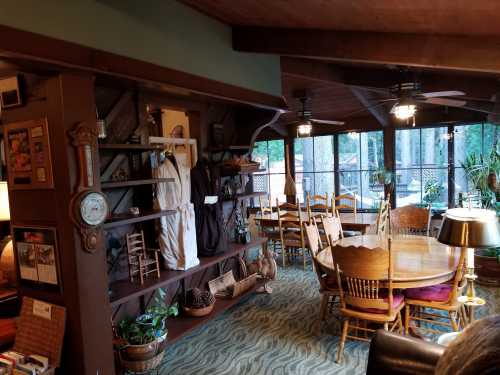 Cozy dining area with wooden furniture, a large table, and a shelf displaying various items, surrounded by windows.