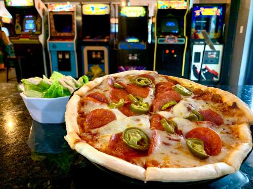 A delicious pepperoni pizza with jalapeños, served with a side salad, in front of colorful arcade machines.