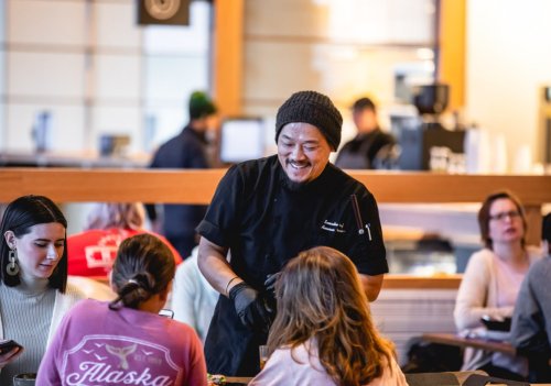 A smiling chef serves food to customers at a bustling restaurant, with diners engaged in conversation.