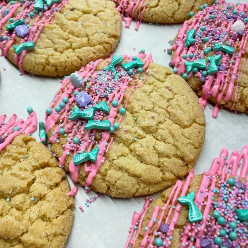 Colorful cookies with pink icing, sprinkles, and decorative mermaid-themed toppings on a white surface.