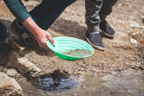 A person using a green pan to sift through sand and gravel near a stream, searching for gold or minerals.
