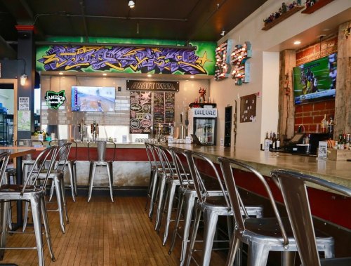 A modern bar interior with metal stools, colorful graffiti art, and screens displaying sports events.