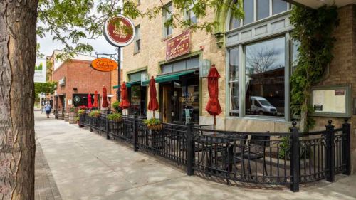 A charming restaurant exterior with outdoor seating, red umbrellas, and greenery along the sidewalk.