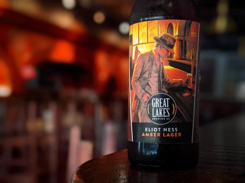 A close-up of a bottle of Great Lakes Eliot Ness Amber Lager, featuring a vintage-style label with a man in a fedora.