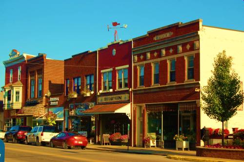 Charming small-town street with colorful storefronts, parked cars, and a clear blue sky.