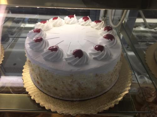 A round cake with white frosting, topped with cherries and decorative swirls, displayed in a glass case.