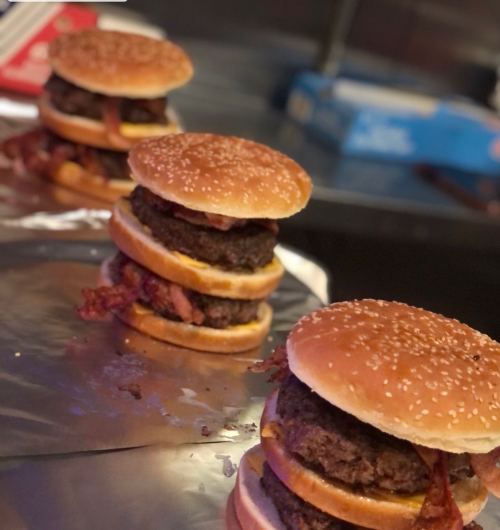 Three stacked burgers with sesame seed buns, featuring layers of beef patties and bacon, on a silver tray.
