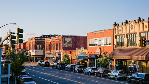 A vibrant downtown street lined with shops, parked cars, and historic buildings under a clear blue sky.