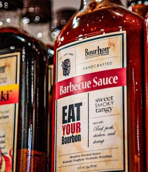Bottles of barbecue sauce on a shelf, featuring a label that reads "Eat Your Bourbon" with a smoky flavor description.