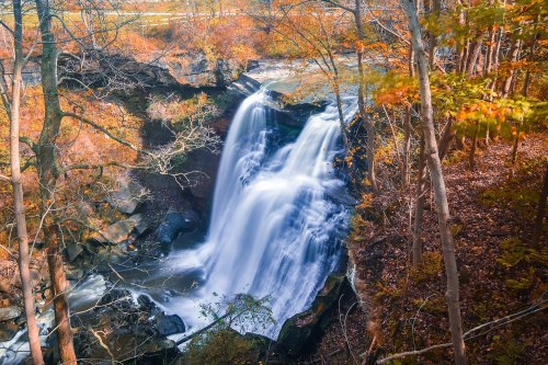 A serene waterfall cascades over rocks, surrounded by vibrant autumn foliage and trees.