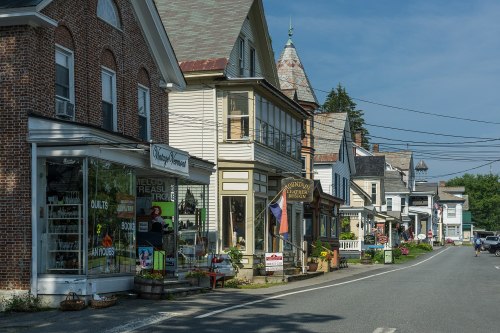 A quaint street lined with historic buildings, shops, and greenery under a clear blue sky.
