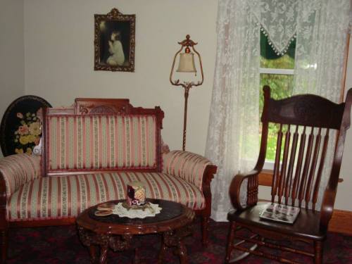 A vintage living room with a striped sofa, wooden rocking chair, ornate table, and lace curtains by a window.