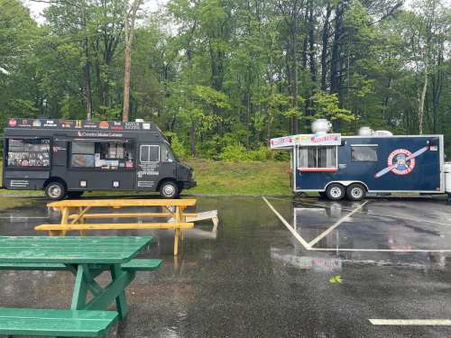 Two food trucks parked in a rainy lot, one black and one silver, with picnic tables nearby.