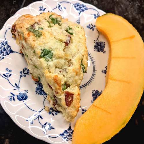 A slice of savory scone with herbs and bacon on a floral plate, accompanied by a wedge of cantaloupe.