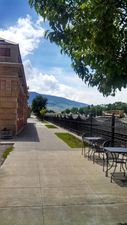 A sunny sidewalk lined with trees, leading to mountains in the distance, with tables and chairs nearby.