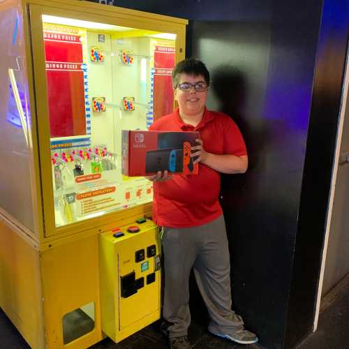 A boy holds a Nintendo Switch in front of a colorful claw machine in an arcade.