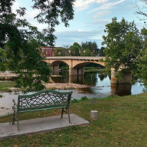 A scenic view of a river with a stone bridge in the background, featuring a bench and lush greenery along the shore.