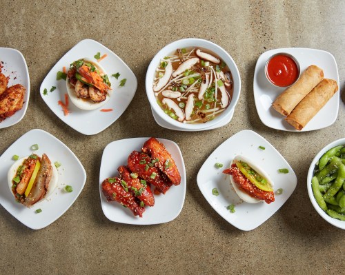A variety of Asian dishes including bao, wings, spring rolls, and soup, arranged on a textured surface.