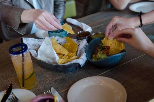 Two hands reaching for tortilla chips from a basket, with guacamole and drinks on a wooden table.