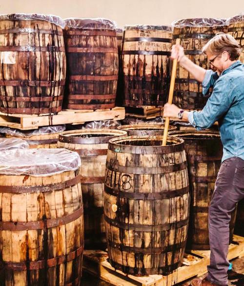 A person stirs a barrel in a warehouse filled with stacked wooden barrels.