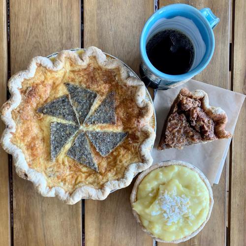 A wooden table displays a pie with a decorative crust, a slice of pie, a small dessert, and a cup of coffee.
