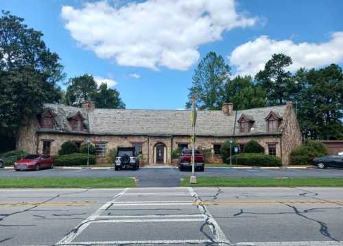 A stone building with a sloped roof, surrounded by trees and parked cars, under a blue sky with clouds.