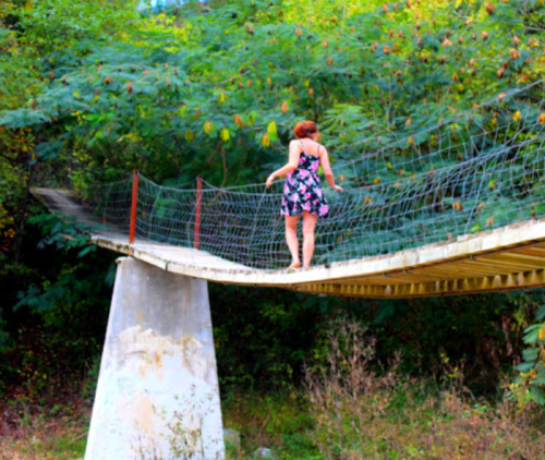 A person in a floral dress walks on a suspended bridge surrounded by lush greenery.