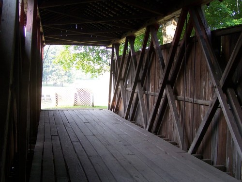 Interior view of a wooden covered bridge, featuring a rustic floor and crisscrossed wooden supports.