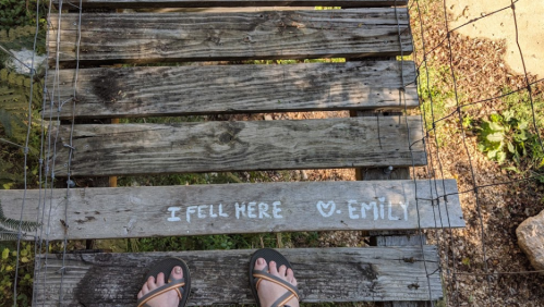 A wooden bridge with a message saying "I fell here ❤️ EMILY" and a pair of feet in sandals on the planks.