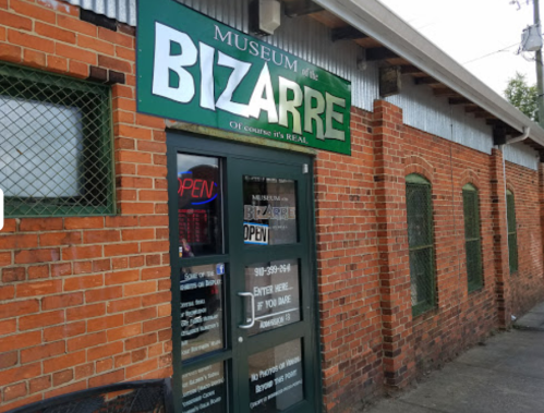 Exterior of the Museum of Bizarre, featuring a green sign and brick walls, with an "Open" notice in the window.