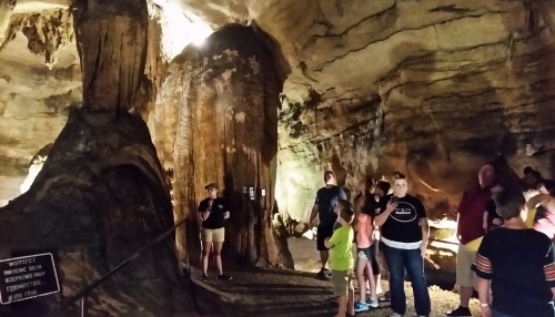 A group of people exploring a cave with rock formations and dim lighting, some reading signs.