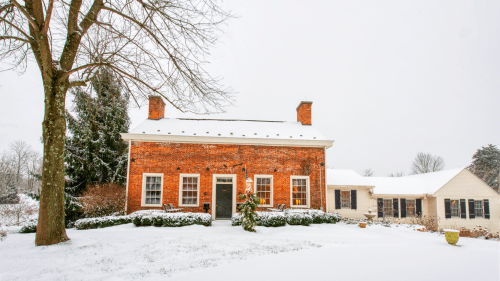 A brick house with a snow-covered roof, surrounded by trees and a snowy yard, creating a cozy winter scene.