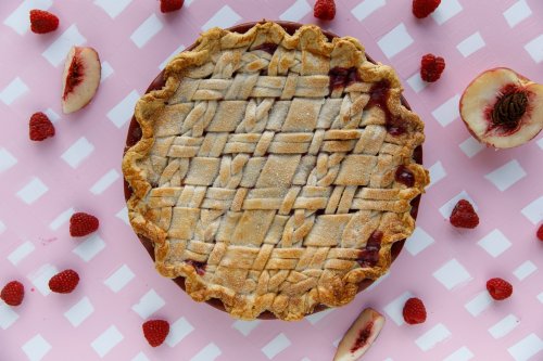 A freshly baked lattice-top pie surrounded by raspberries and peach slices on a pink checkered background.