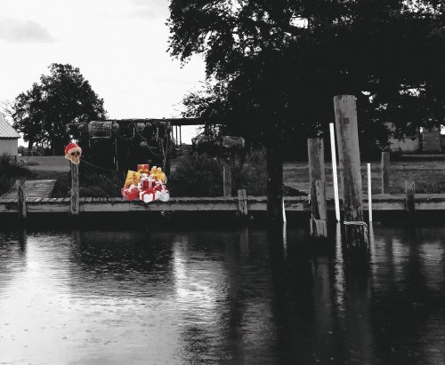 A black-and-white scene of a riverbank with a colorful Santa and gifts on a dock, surrounded by trees.