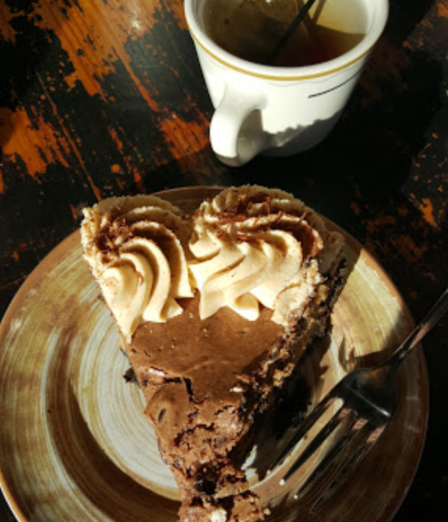 A slice of chocolate cake with cream frosting on a plate, next to a cup of coffee on a wooden table.