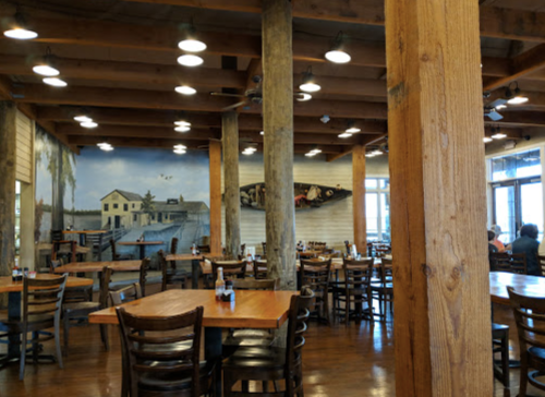 Interior of a rustic restaurant with wooden beams, tables, and a mural of a waterfront scene on the wall.