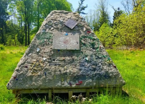 A large, weathered stone monument covered in colorful markings, surrounded by green grass and trees in a sunny landscape.