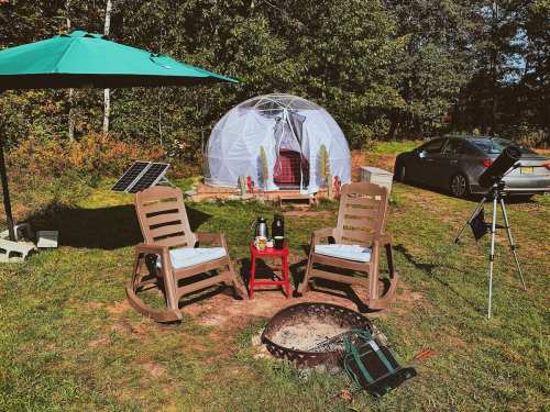 A cozy outdoor setup with two rocking chairs, a fire pit, a dome tent, and a car nearby, surrounded by trees.
