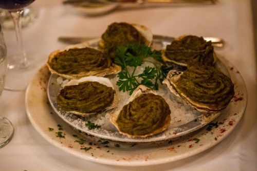 A plate of baked oysters topped with a green herb mixture, garnished with parsley, on a bed of ice.
