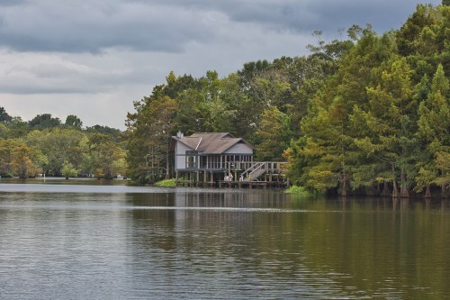 A serene lake scene featuring a wooden house on stilts surrounded by lush trees under a cloudy sky.
