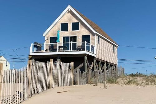 A beach house on stilts with a deck, surrounded by sand and wooden fencing, under a clear blue sky.