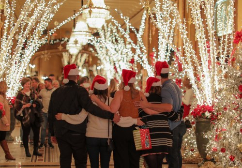 A group of people wearing Santa hats stands together, surrounded by festive lights and decorations in a holiday setting.