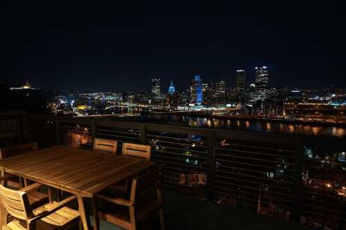A nighttime cityscape view of a skyline with illuminated buildings and a river, featuring a wooden table and chairs in the foreground.